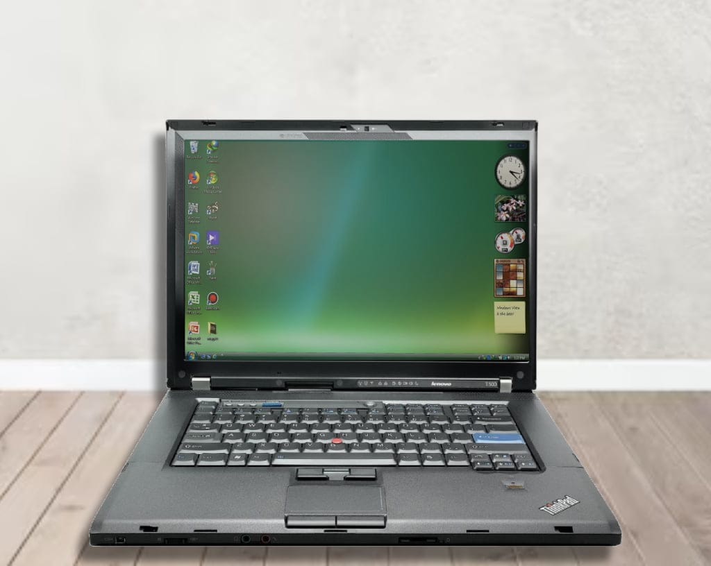 A Lenovo ThinkPad T500 laptop computer sitting on a wooden floor. The laptop has a black exterior and a silver keyboard.